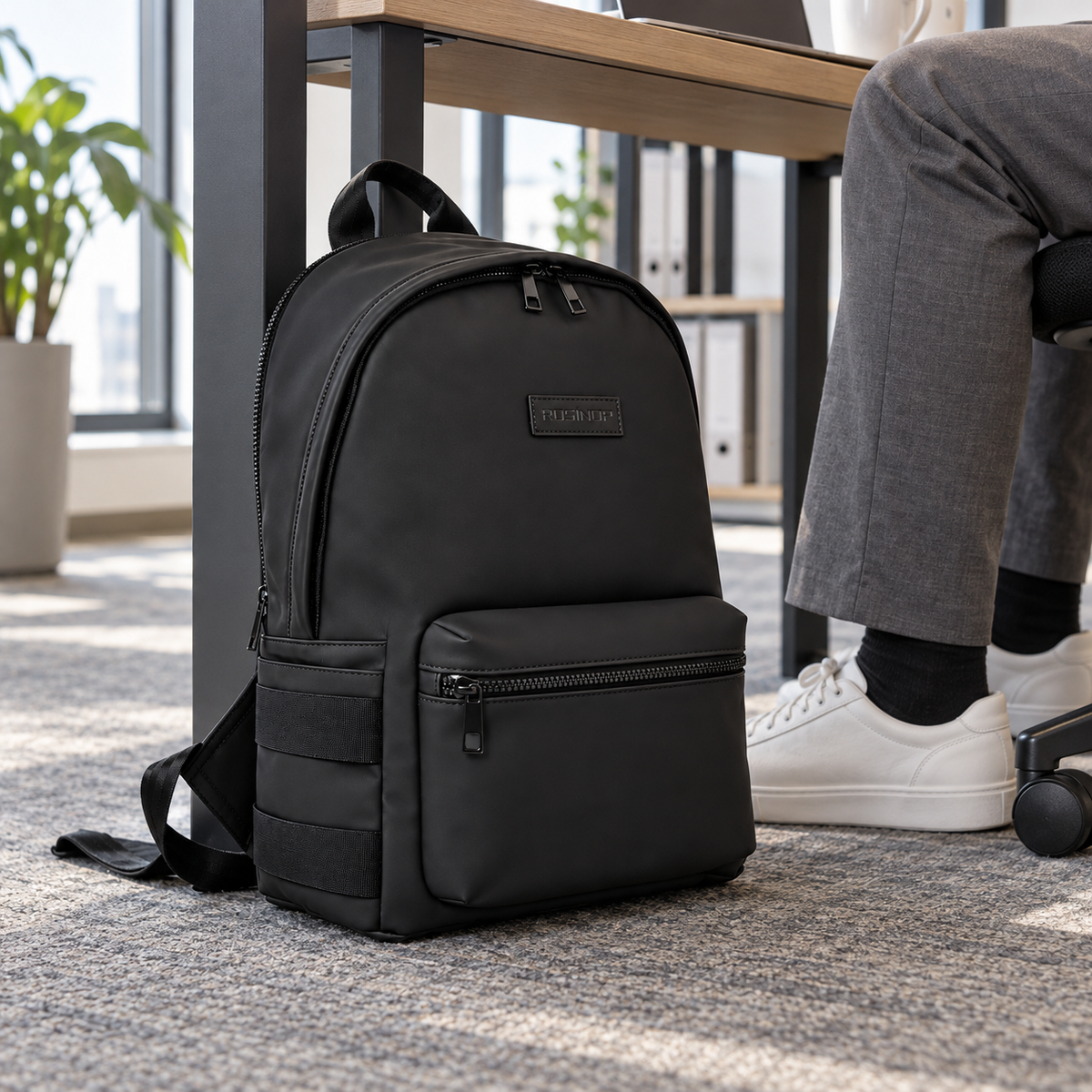 Black ROSINOP backpack resting beside a desk during an ordinary workday.